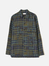 Olive shadows check fatigue shirt laid flat on a light grey seamless backdrop. The shirt features a camp collar, a single patch chest pocket with a buttoned flap, and a five-button placket with dark buttons. Long sleeves with plain cuffs. The fabric is a patchwork-style plaid in olive, navy, and teal with a textured weave.