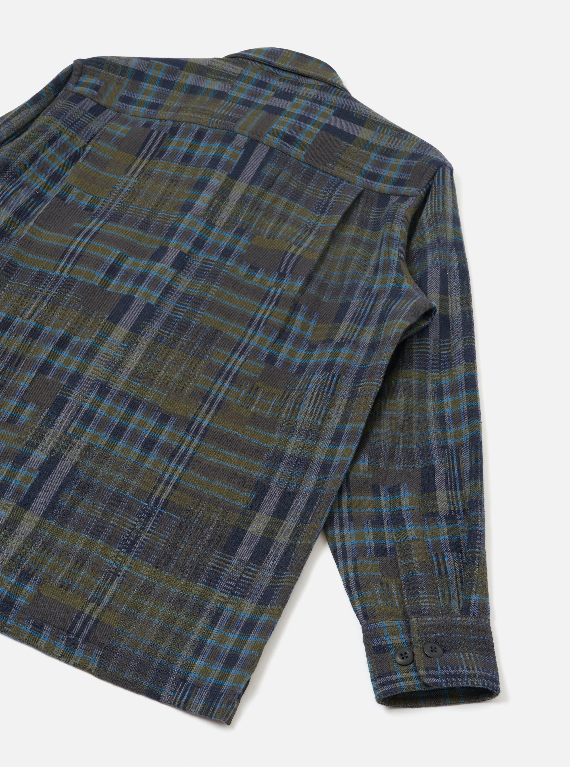 Flat lay of an olive shadows check fatigue shirt on a white studio surface, shot from above at an angle. The back panel and one long sleeve are visible, showing a brushed flannel fabric in an olive, navy, grey and blue check pattern. The buttoned cuff features two dark buttons.