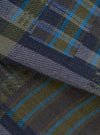 Close-up flat lay of the olive shadows check fabric used in the fatigue shirt. The woven textile shows a tartan-style check in olive green, navy and slate blue, with bright cyan overcheck stripes. A diagonal fold runs across the centre, revealing the twill weave texture in detail.