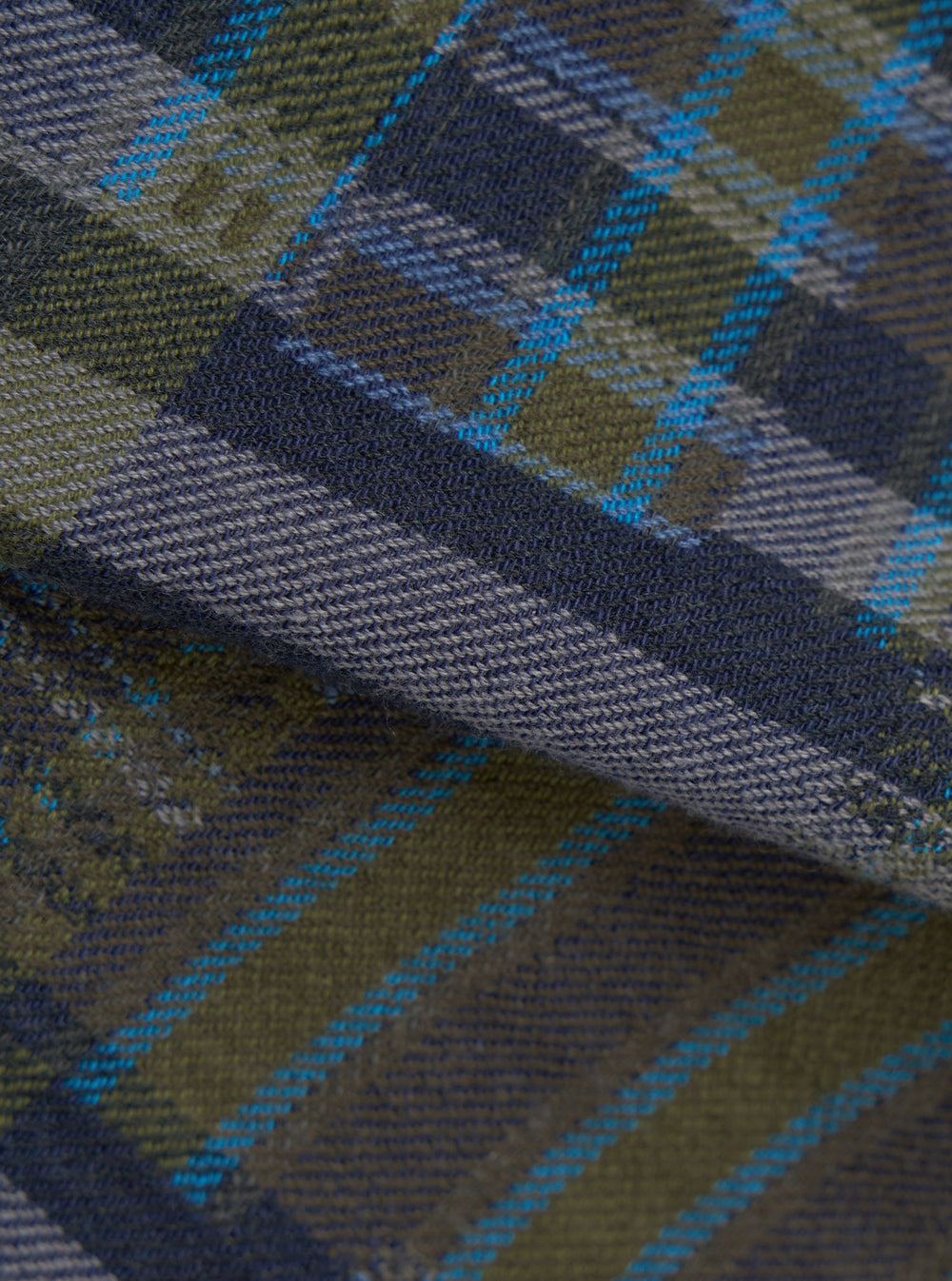 Close-up flat lay of the olive shadows check fabric used in the fatigue shirt. The woven textile shows a tartan-style check in olive green, navy and slate blue, with bright cyan overcheck stripes. A diagonal fold runs across the centre, revealing the twill weave texture in detail.