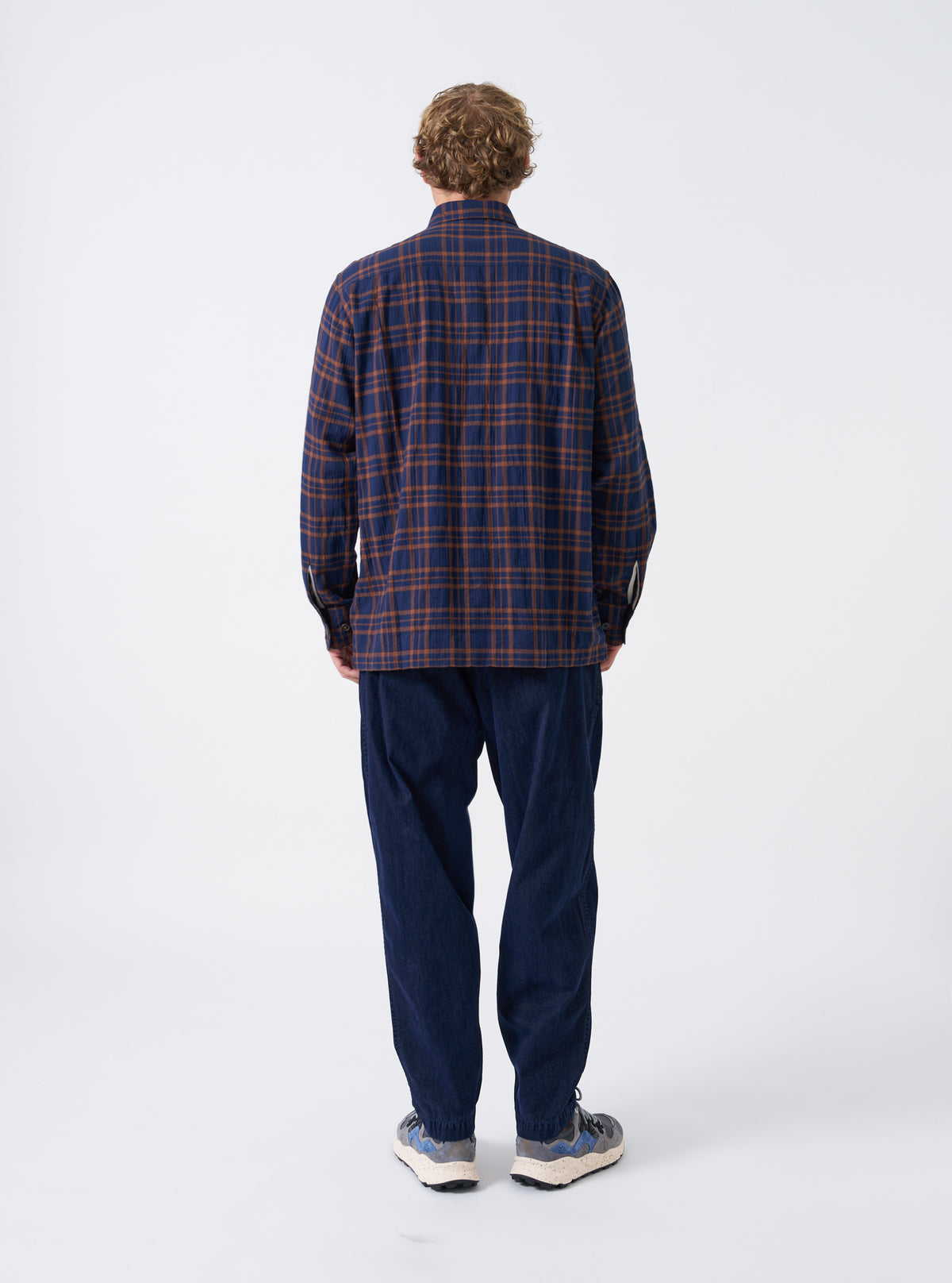 A back view of a model wearing the Navy Flock Chambray Pleated Track Pant against a plain background. The image highlights the rear pockets, relaxed cut, and dark blue flock chambray cotton fabric draped down the legs.