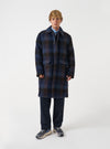 A model wearing the Universal Works Navy/Brown Berlin Wool Check Account Coat, shown from the front against a plain studio background. The model wears the loose-fitting checkered wool coat buttoned up.