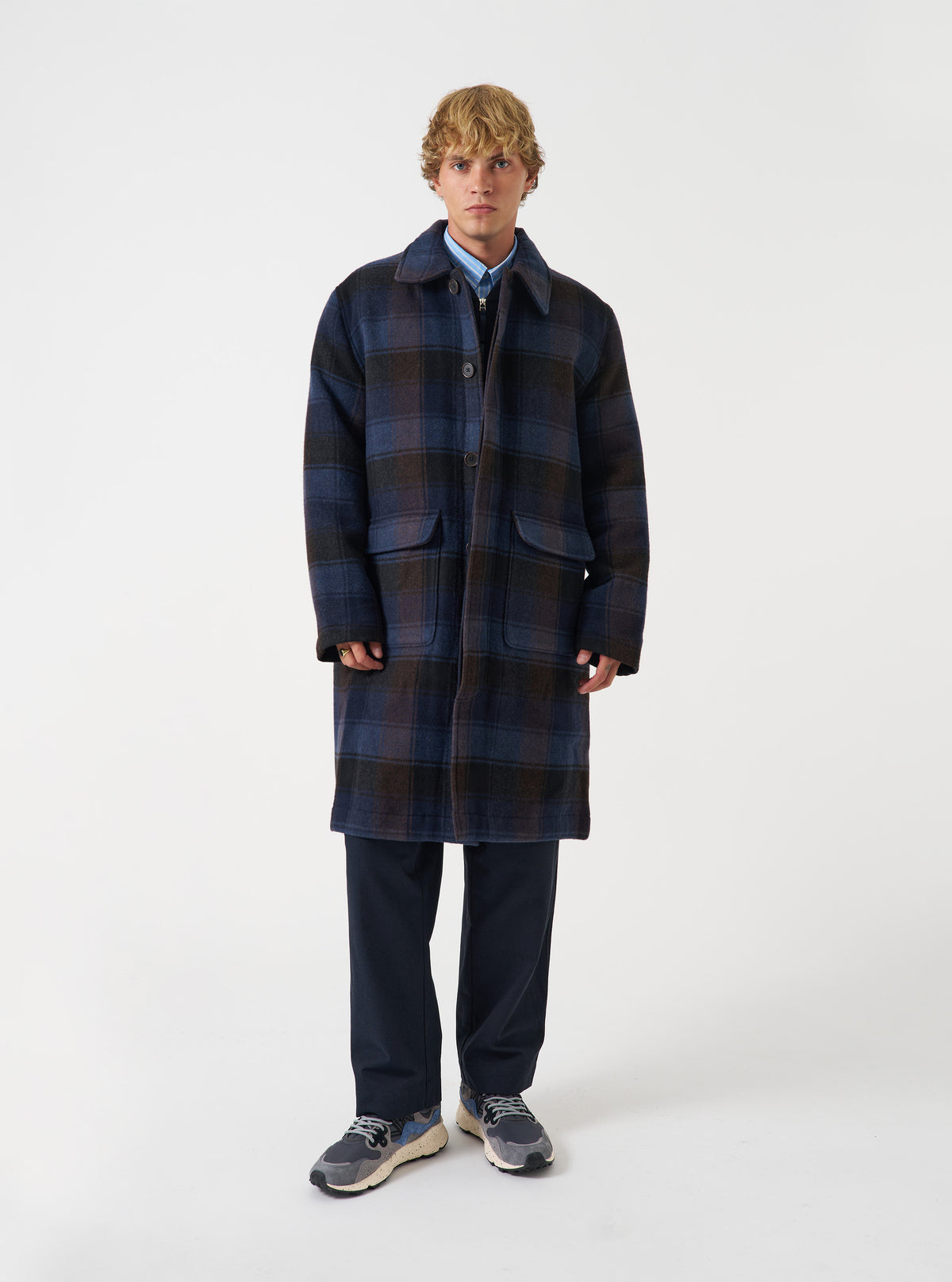 A model wearing the Universal Works Navy/Brown Berlin Wool Check Account Coat, shown from the front against a plain studio background. The model wears the loose-fitting checkered wool coat buttoned up.