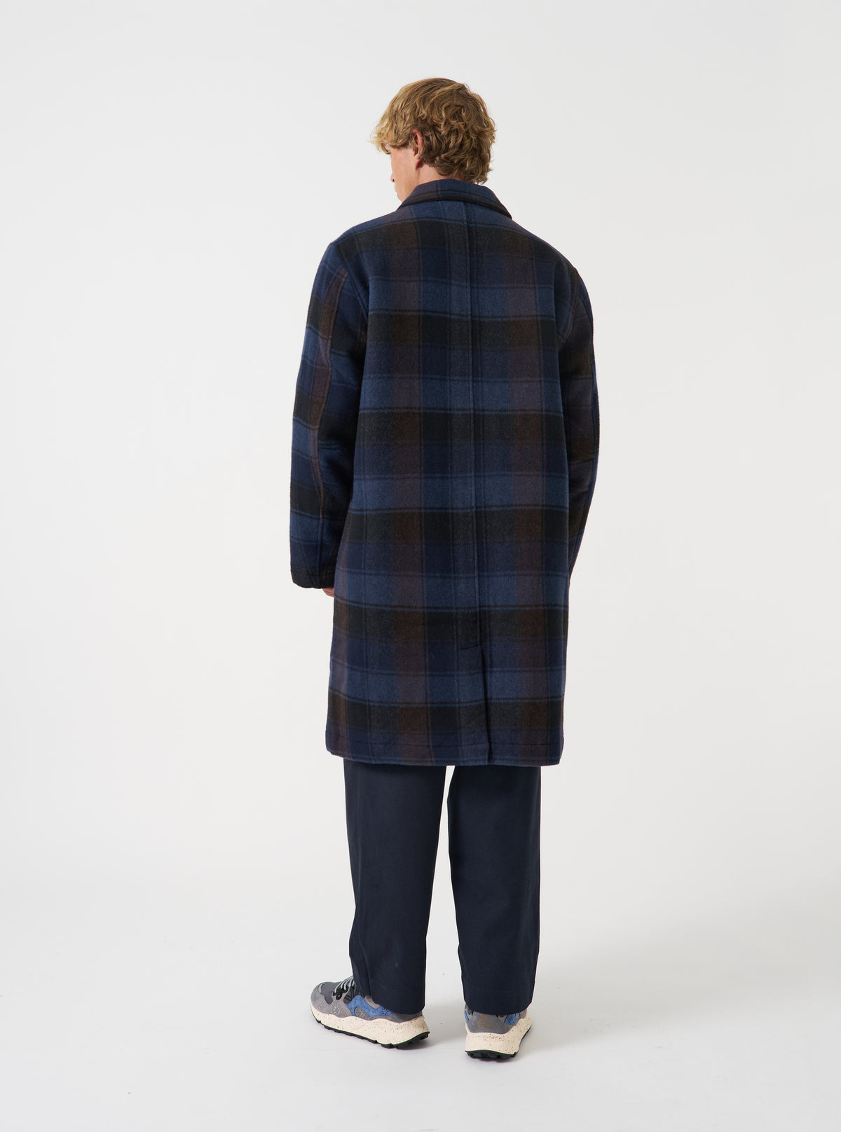 A back view of a model wearing the Universal Works Navy/Brown Berlin Wool Check Account Coat against a plain studio backdrop, displaying the single rear vent and oversized fit across the shoulders.