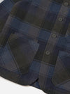 A detailed close-up shot of the Universal Works Navy/Brown Berlin Wool Check OG Bakers Chore Jacket on a plain background, highlighting the signature angled top buttonhole and classic collar details.