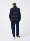 A back view of a model wearing the Universal Works Navy/Brown Berlin Wool Check OG Bakers Chore Jacket set against a plain backdrop, showing the overall fit, straight hem, and center back seam construction.