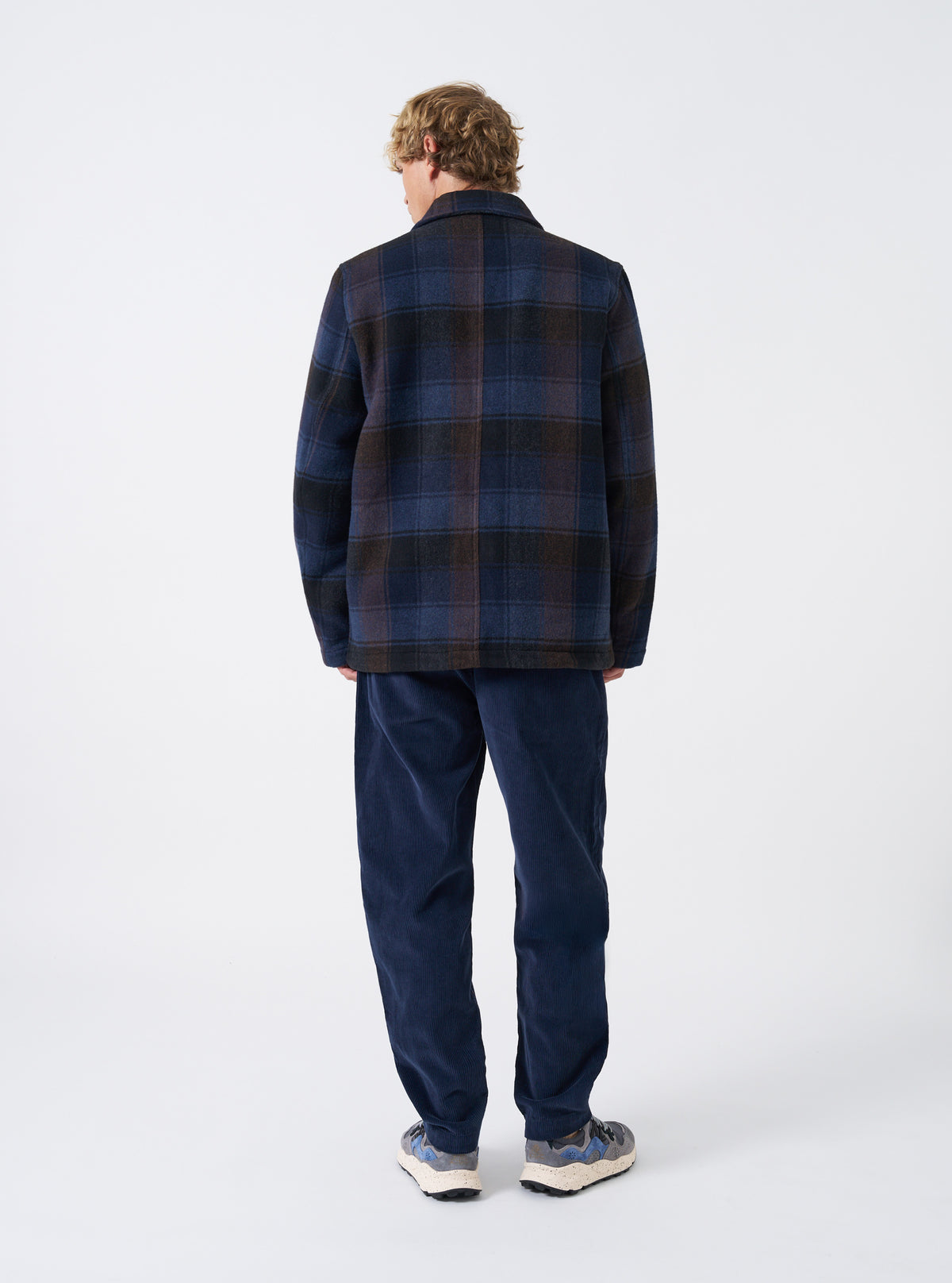A back view of a model wearing the Universal Works Navy/Brown Berlin Wool Check OG Bakers Chore Jacket set against a plain backdrop, showing the overall fit, straight hem, and center back seam construction.