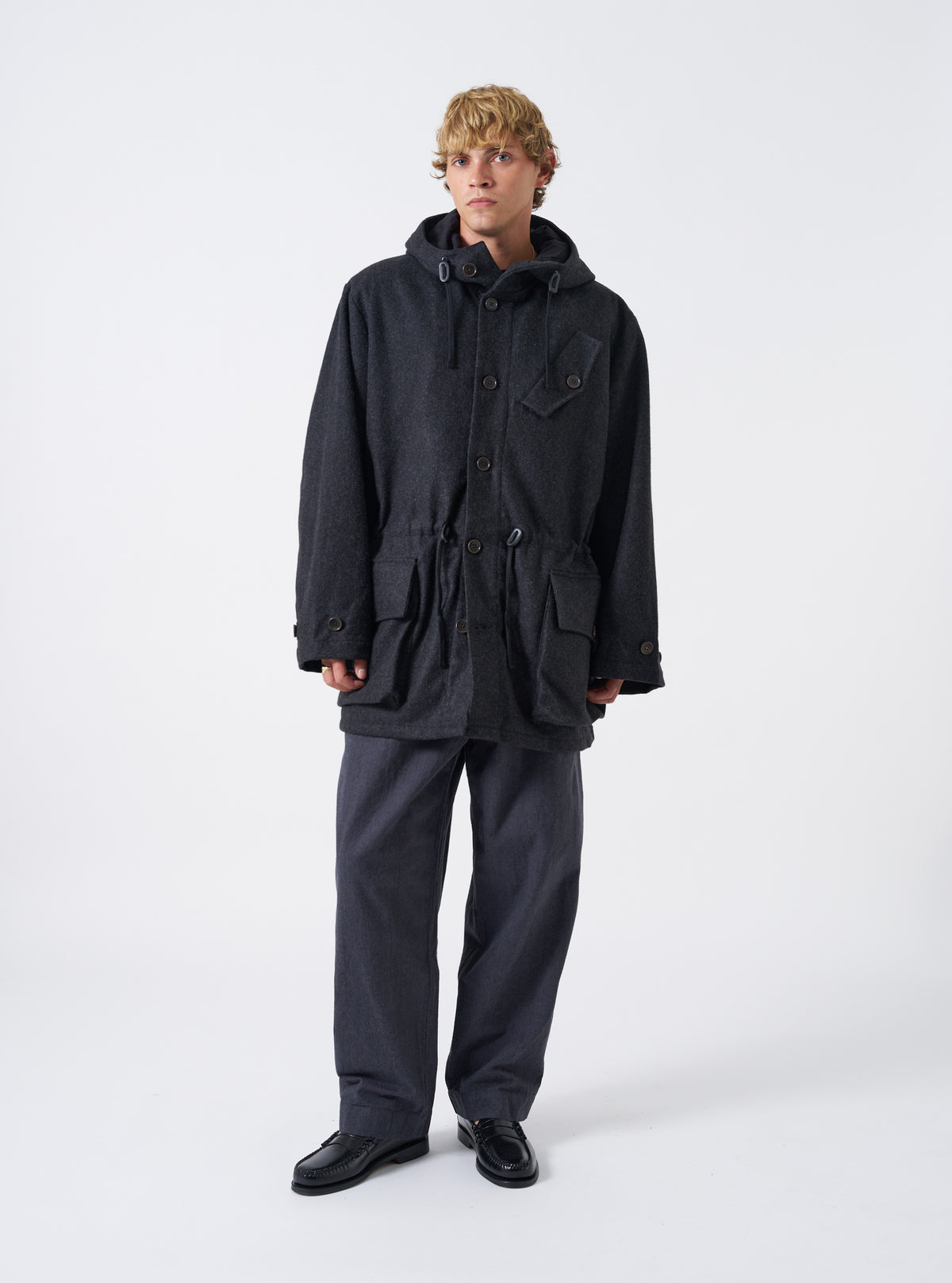 A model wearing the Universal Works Grey Marl Mowbray Boston Parka, captured from the front on a plain backdrop. The grey wool-blend hooded parka displays prominent 3D front pockets and a diagonal chest pocket.