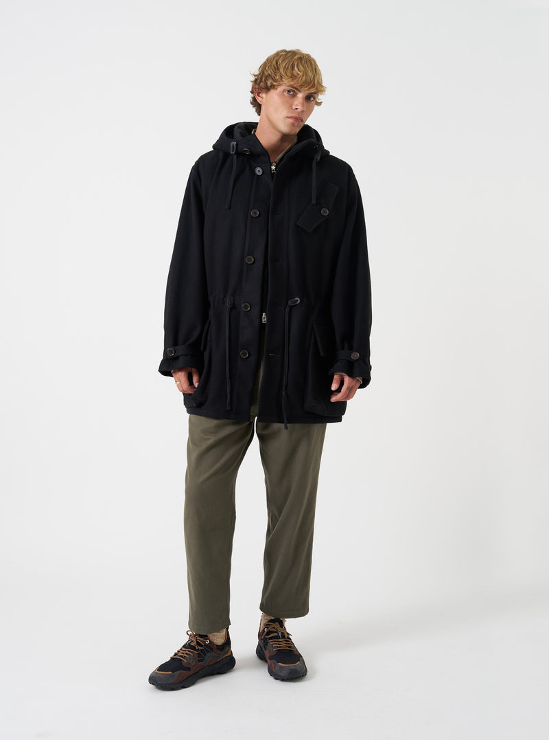 A model wearing the Universal Works Black Mowbray Boston Parka, shown from the front against a plain background. The black wool-blend parka features a spacious hood, front button closure, and large 3D patch pockets.