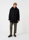 A model wearing the Universal Works Black Mowbray Boston Parka, shown from the front against a plain background. The black wool-blend parka features a spacious hood, front button closure, and large 3D patch pockets.