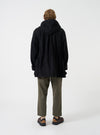 A back view of a model wearing the Universal Works Black Mowbray Boston Parka against a plain studio backdrop, highlighting the oversized silhouette, deep hood, adjustable waist cinch, and mid-length cut.