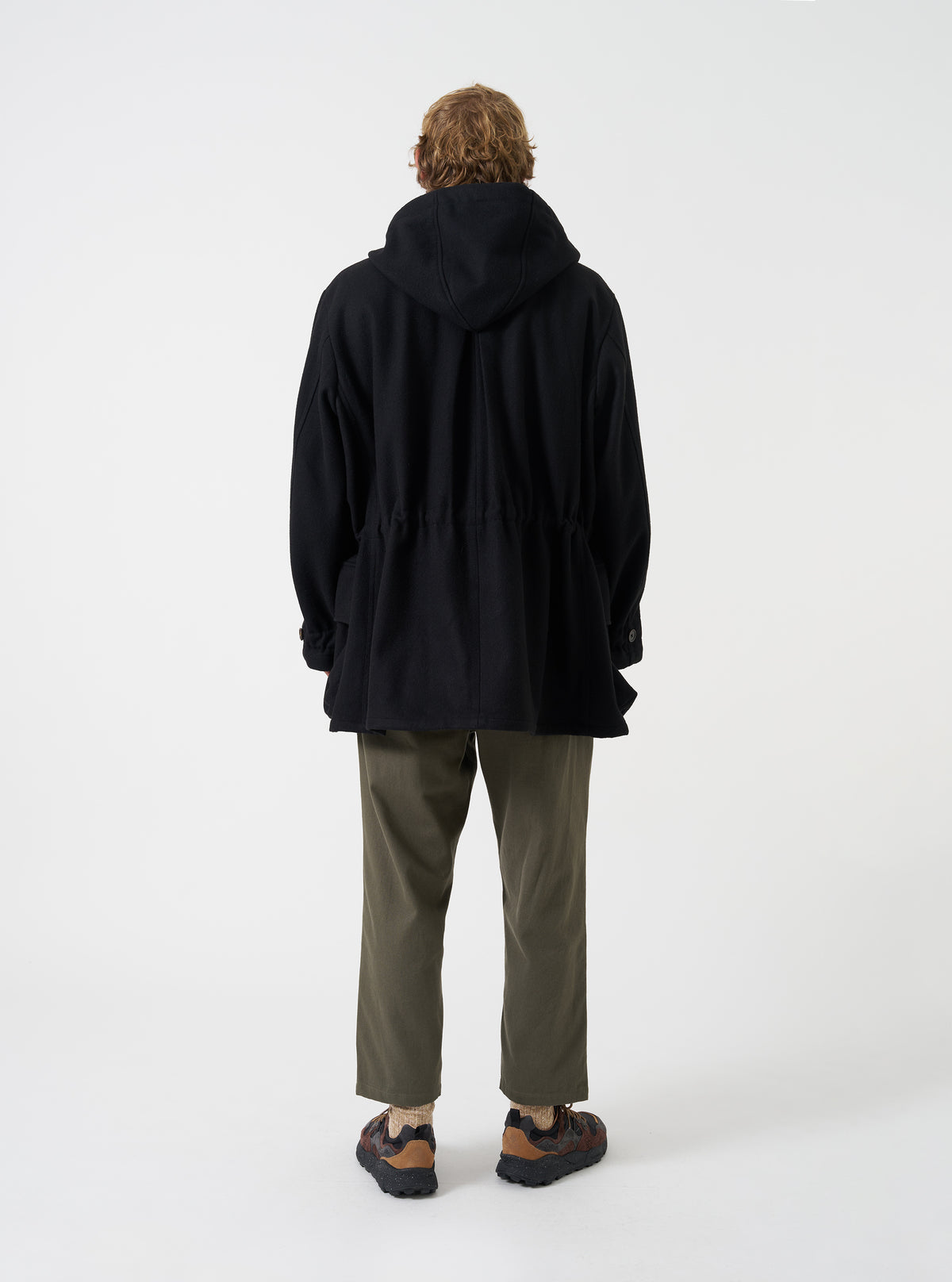 A back view of a model wearing the Universal Works Black Mowbray Boston Parka against a plain studio backdrop, highlighting the oversized silhouette, deep hood, adjustable waist cinch, and mid-length cut.