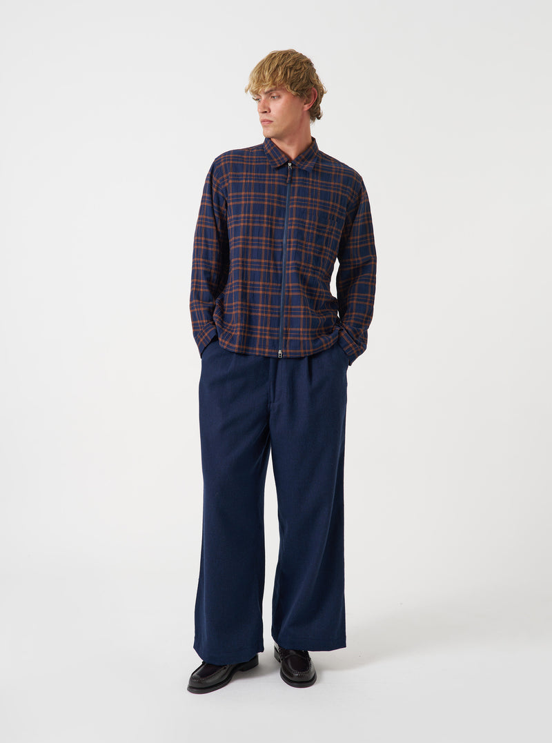 A front view of a model wearing the Universal Works Navy/Brown Young Check Zip Shirt. The relaxed-fit overshirt features a full zip front, classic pointed collar, straight front hem, and a single chest patch pocket against a plain backdrop.