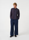 A front view of a model wearing the Universal Works Navy/Brown Young Check Zip Shirt. The relaxed-fit overshirt features a full zip front, classic pointed collar, straight front hem, and a single chest patch pocket against a plain backdrop.