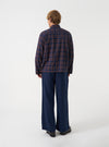 A side view of a model wearing the Universal Works Navy/Brown Young Check Zip Shirt over a plain t-shirt. The image highlights the relaxed silhouette, dropped shoulder seams, straight cuffs, and subtle rear hem curve against a neutral backdrop.