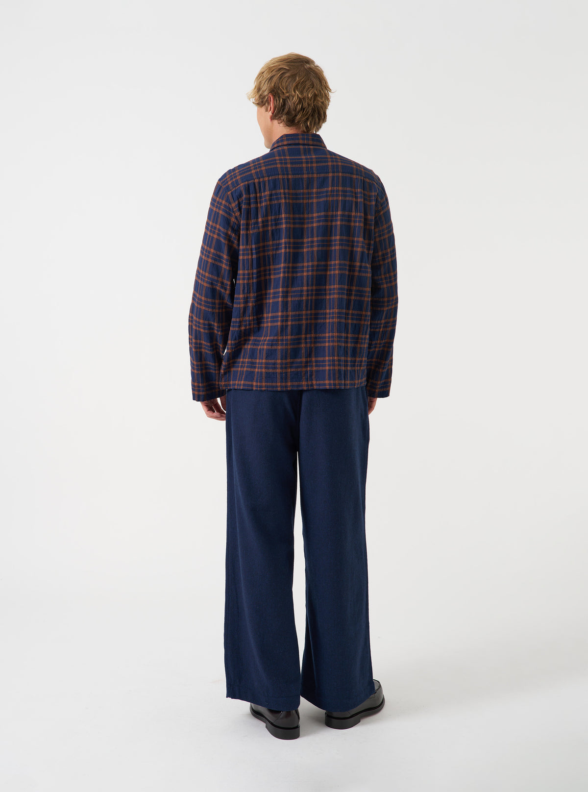 A side view of a model wearing the Universal Works Navy/Brown Young Check Zip Shirt over a plain t-shirt. The image highlights the relaxed silhouette, dropped shoulder seams, straight cuffs, and subtle rear hem curve against a neutral backdrop.