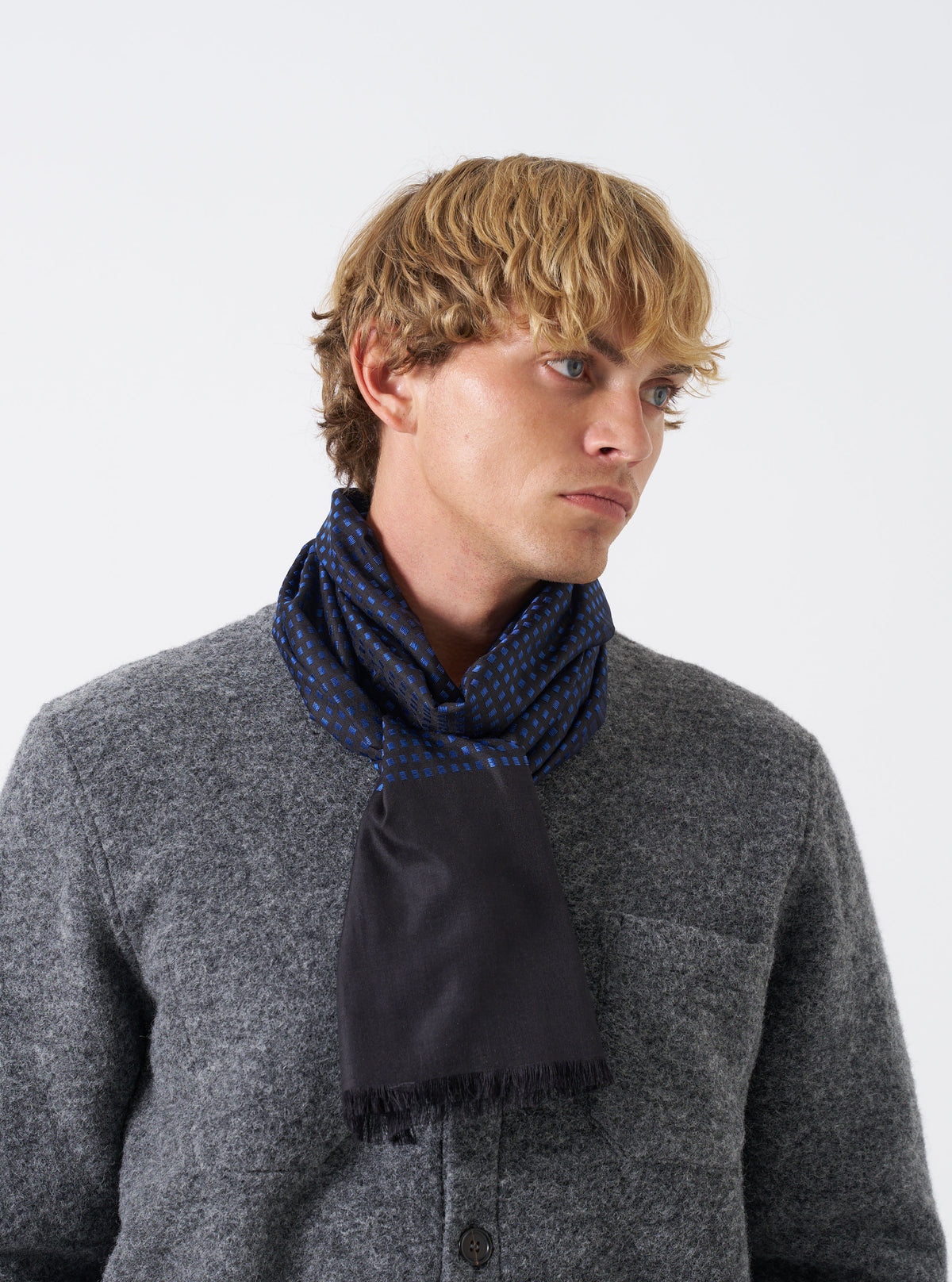 A model wearing the Universal Works Black/Blue Jacquard Silk Scarf wrapped around the neck. The lightweight silk accessory features a woven black and blue pattern, smooth finish, and elegant drape set against a plain neutral background.