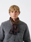 A model wearing the Universal Works Brown/Sky/Gold Jacquard Silk Scarf styled around the neck. The lightweight silk scarf showcases an intricate woven geometric pattern in brown, sky blue, and gold tones set against a plain neutral backdrop.