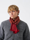 A model wearing the Universal Works Red/Silver/Gold Jacquard Silk Scarf wrapped comfortably around the neck. The pure silk scarf features a multi-coloured jacquard weave with vibrant red, silver, and gold accents against a plain background.