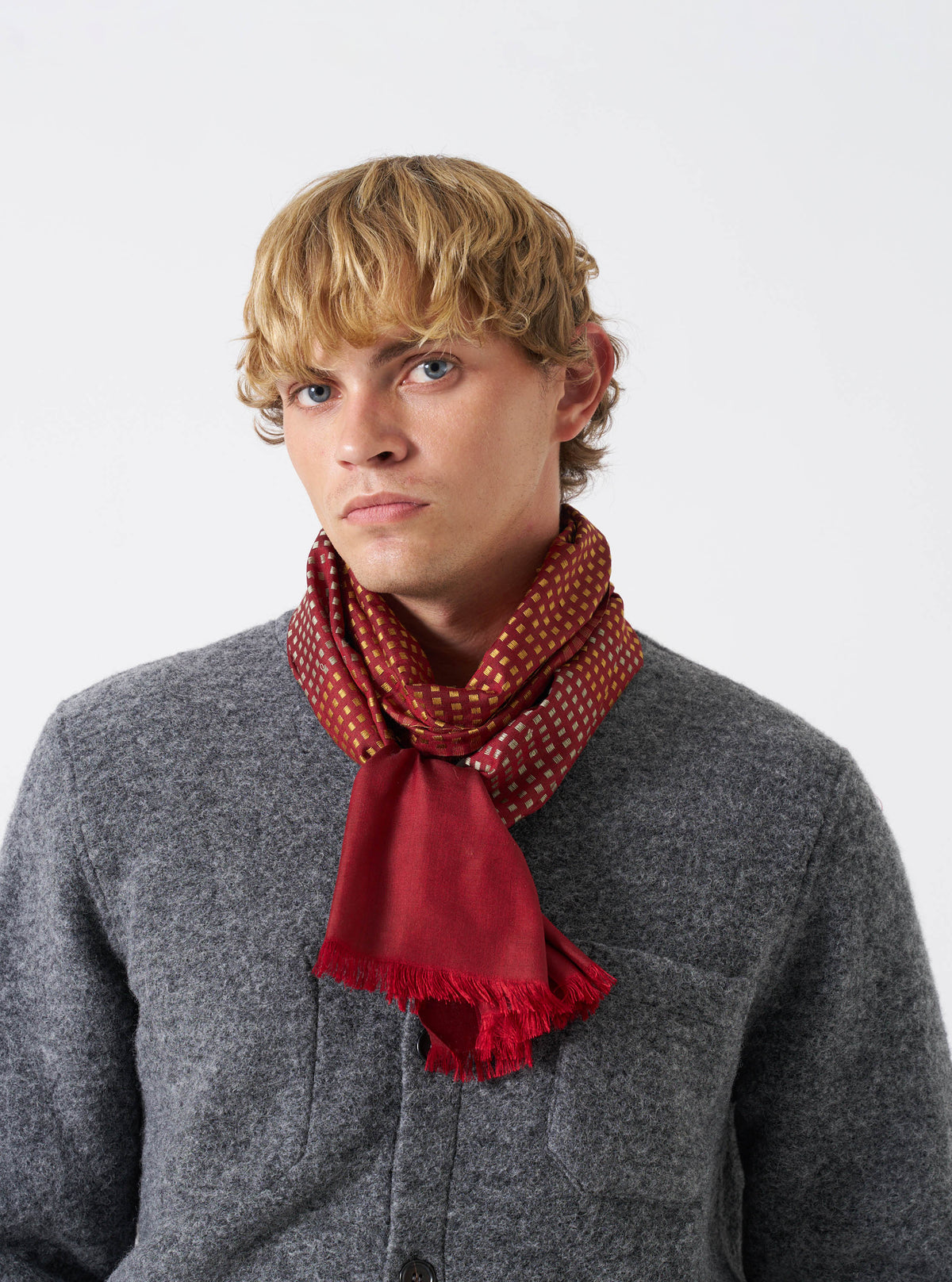 A model wearing the Universal Works Red/Silver/Gold Jacquard Silk Scarf wrapped comfortably around the neck. The pure silk scarf features a multi-coloured jacquard weave with vibrant red, silver, and gold accents against a plain background.