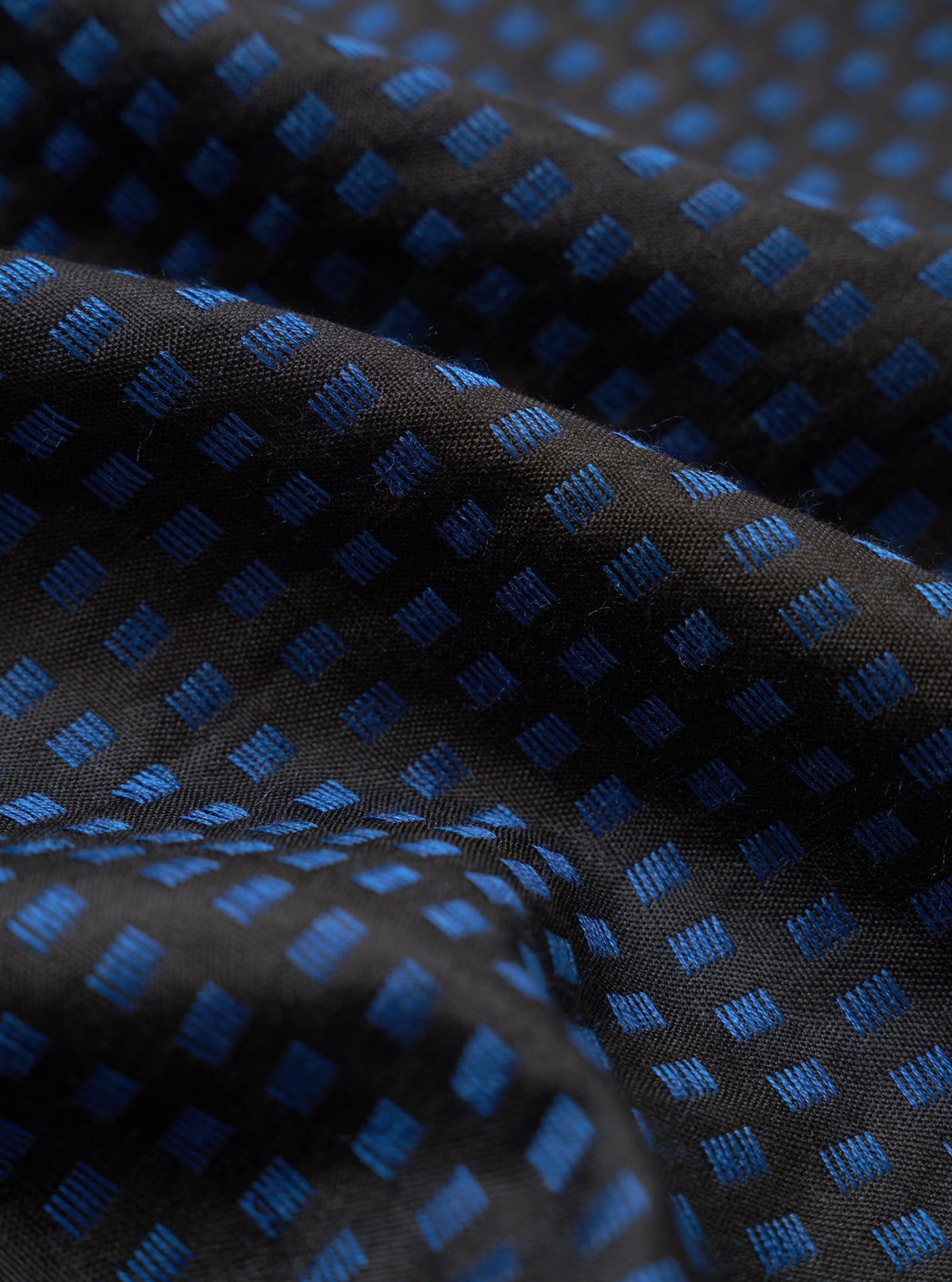 A close-up swatch detail of the Universal Works Black/Blue Jacquard Silk Scarf. The extreme close-up view emphasizes the intricate weave of the black and blue silk jacquard fabric and its fine thread structure against a plain background.