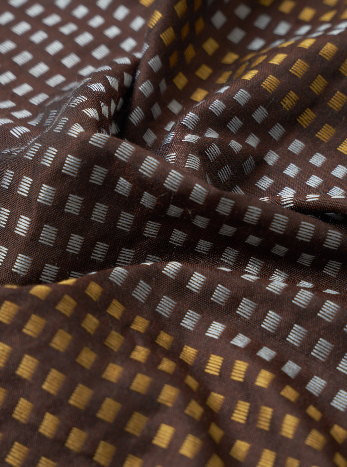 A close-up macro image of the Universal Works Brown/Sky/Gold Jacquard Silk Scarf fabric swatch. The detailed image showcases the texture of the pure silk jacquard weave and its distinct colour pattern.