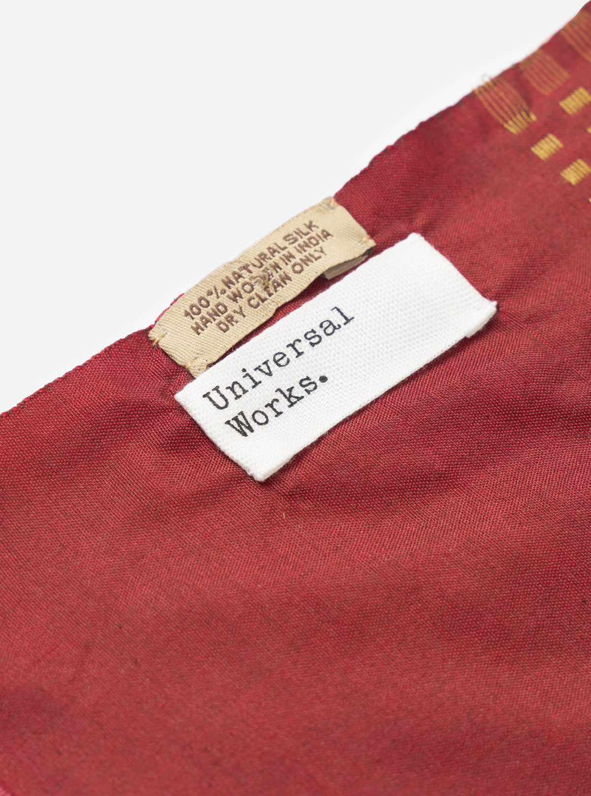 A draped view of the Universal Works Red/Silver/Gold Jacquard Silk Scarf showcasing the soft folds of the silk material. The red, silver, and gold patterned accessory is displayed against a clean background.