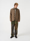A model wearing the Universal Works Sand/Brown Harris Tweed OG Bakers Chore Jacket from the front. The loose-fit jacket features a classic collar, front button closure, signature angled patch pockets, and heavy tweed fabric against a plain backdrop.