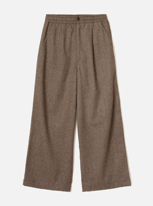 Brown wool herringbone wide-leg trousers laid flat on a plain white background. The elasticated waistband features a single dark button at centre front. Slanted side pockets sit below the waistband. The fabric shows a classic herringbone weave in brown and cream tones. Full-length legs with a generous wide leg and plain hem.