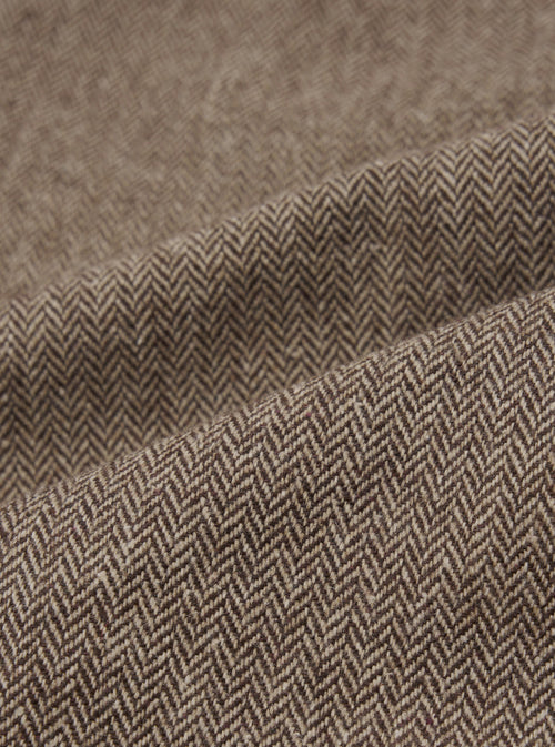 Close-up detail shot of the brown wool herringbone fabric used in the Gillespie Pant. The weave shows a distinct V-shaped herringbone pattern in dark brown and ecru tones. The fabric is folded to reveal texture and weight, with a slightly flecked, woollen surface visible across the folds.
