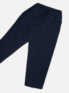 The Navy Wool Herringbone Pleated Track Pant is laid flat on a light grey surface, displayed from the back. Highlights include an elasticated waistband, two buttoned rear welt pockets, fine herringbone wool fabric, and legs tapering to plain hems.
