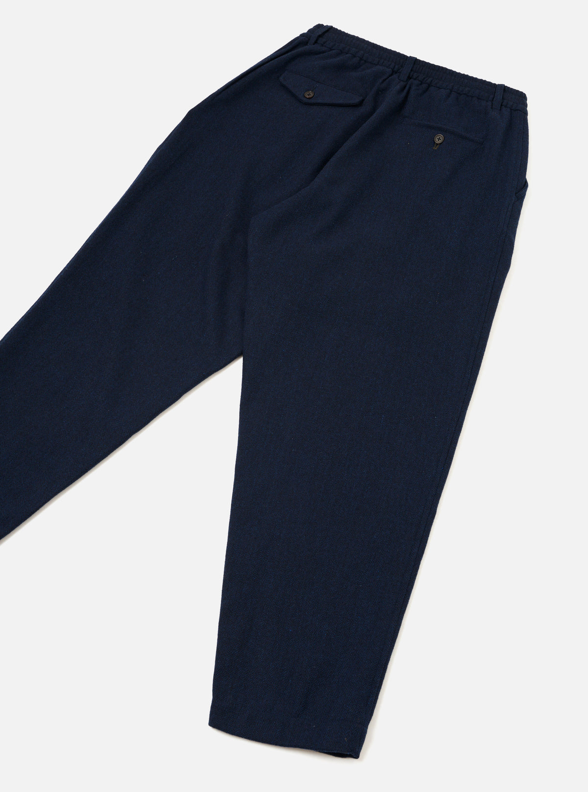 The Navy Wool Herringbone Pleated Track Pant is laid flat on a light grey surface, displayed from the back. Highlights include an elasticated waistband, two buttoned rear welt pockets, fine herringbone wool fabric, and legs tapering to plain hems.