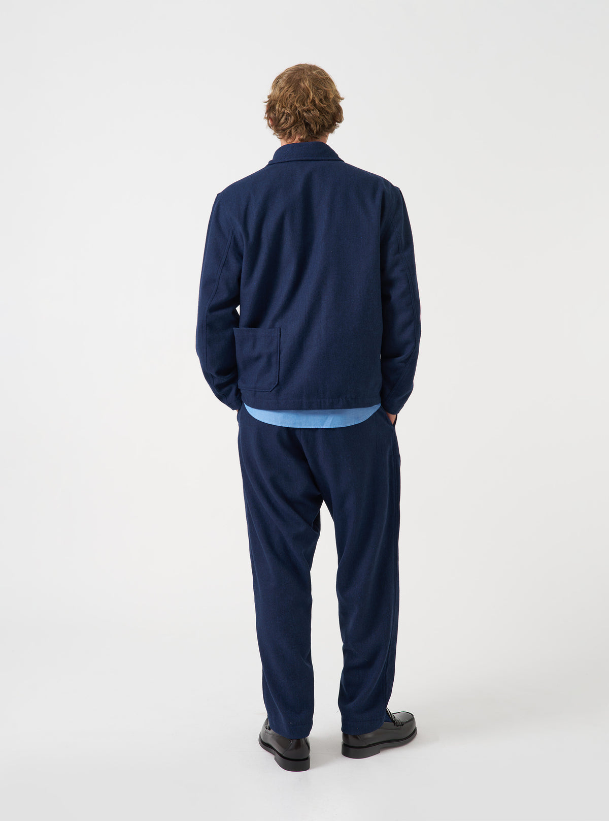 A back view of a model wearing the Navy Wool Herringbone Ives Jacket. The image focuses on the rear fit of the navy wool-blend jacket, highlighting the clean stitching, relaxed shoulders, and straight hem against a plain backdrop.