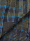 Close-up of the olive shadows check fabric used in the Franck Jacket. The woven twill shows a large-scale tartan check in olive green and navy, with accent stripes in bright teal and grey. The fabric is folded to reveal texture and drape.