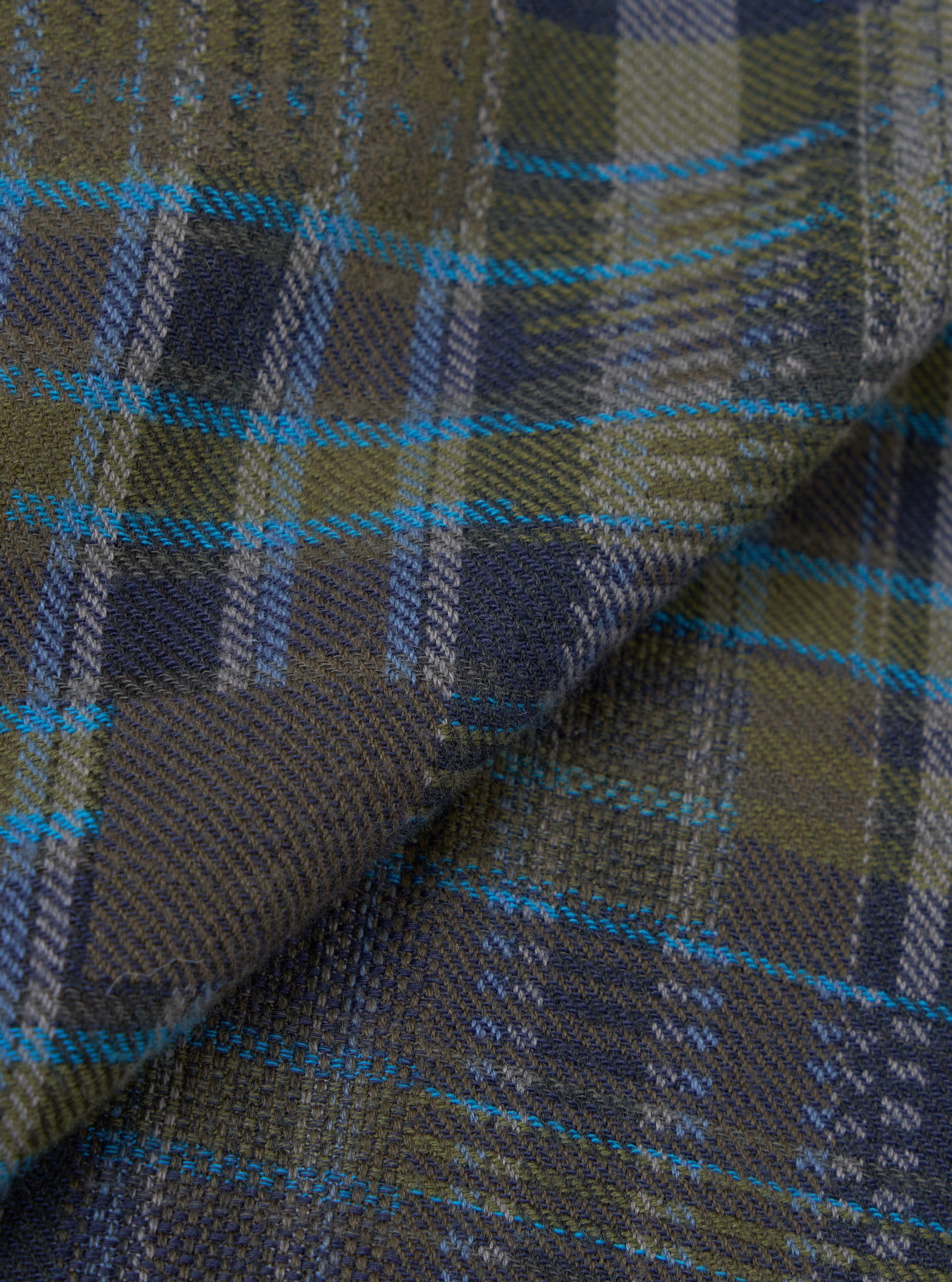 Close-up of the olive shadows check fabric used in the Franck Jacket. The woven twill shows a large-scale tartan check in olive green and navy, with accent stripes in bright teal and grey. The fabric is folded to reveal texture and drape.