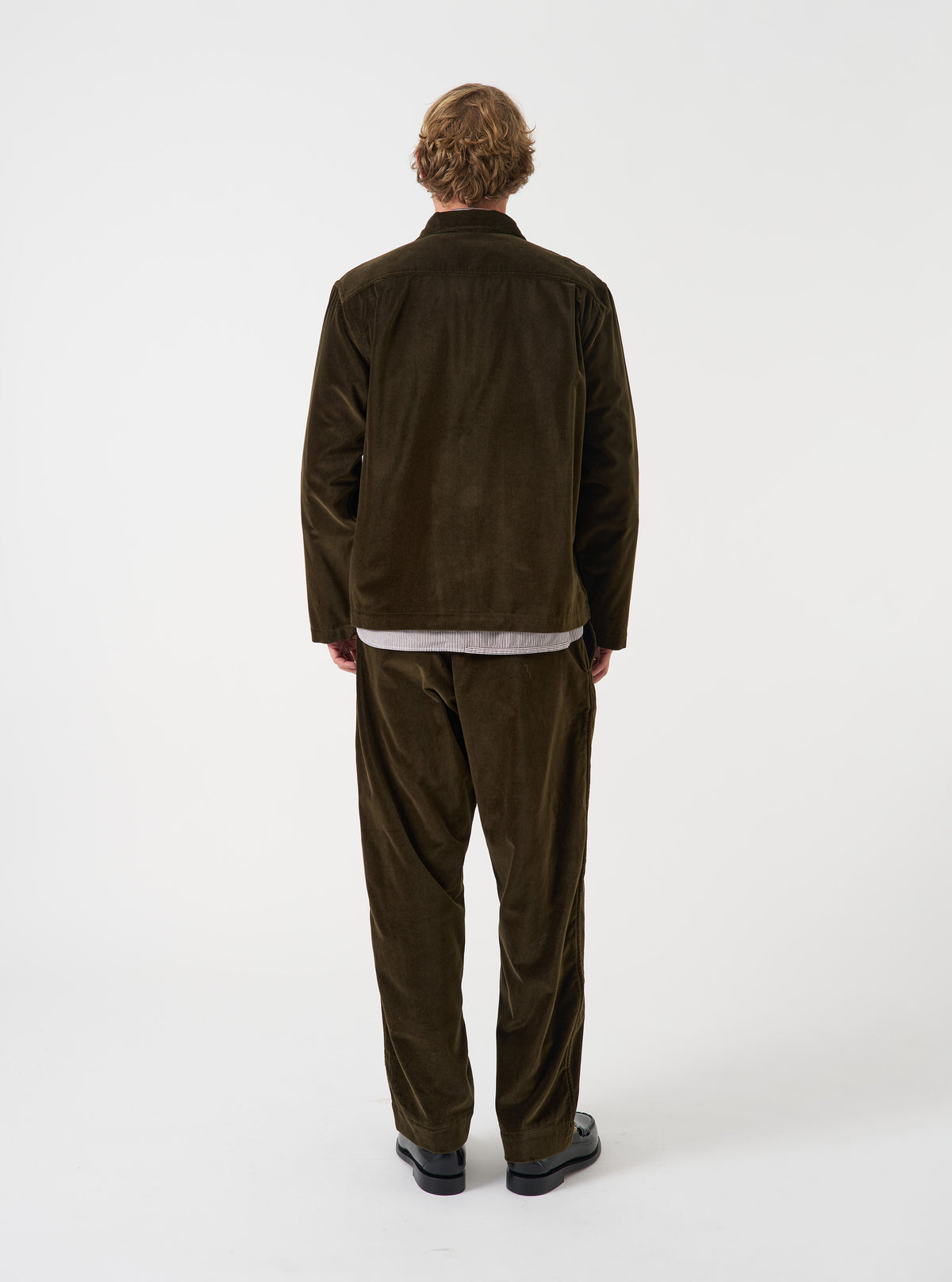 A back view of a model wearing the Olive CPH Velvet Zip OS Shirt. The photograph showcases the back panel of the olive green cotton velvet overshirt, emphasizing its relaxed silhouette and straight hem against a plain backdrop.