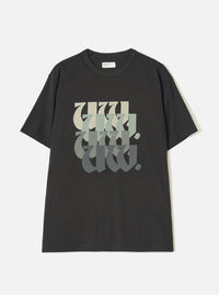 Black single jersey t-shirt laid flat on a white studio surface. Crew neck with a small Universal Works woven label at the back neck. Short sleeves with a relaxed fit. Large chest graphic featuring stacked "UW" lettering in a blackletter style, fading from cream through sage green to dark grey.