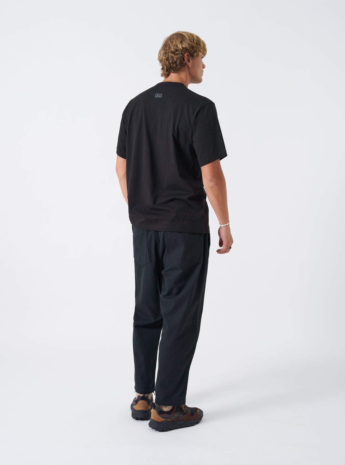 A back view of a model wearing the Universal Works Black Single Jersey UW Print Core Tee. The image showcases the smooth fit across the shoulders and the clean straight hemline of the black cotton t-shirt against a plain background.