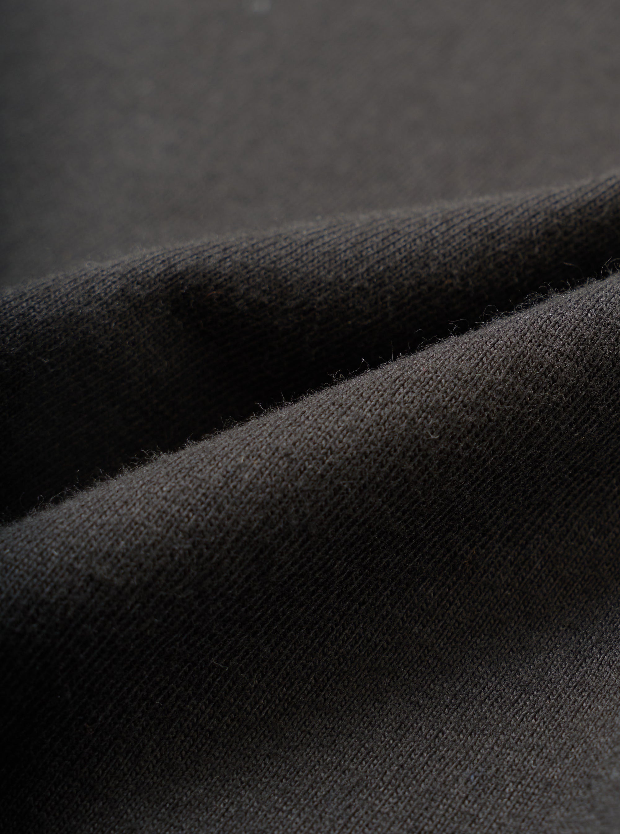 Close-up detail shot of the black single jersey fabric used in the Core Tee, showing a fine knit texture with a soft, slightly raised surface. The material is folded to reveal its weight and drape. Shot against a dark grey seamless backdrop.