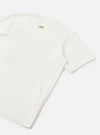 Flat lay of an ecru single jersey t-shirt, shot from above at an angle showing the back. A small olive green branded logo print sits at the upper back centre. Short sleeves with ribbed cuffs are visible. The fabric has a smooth, fine jersey texture. Plain white studio surface.