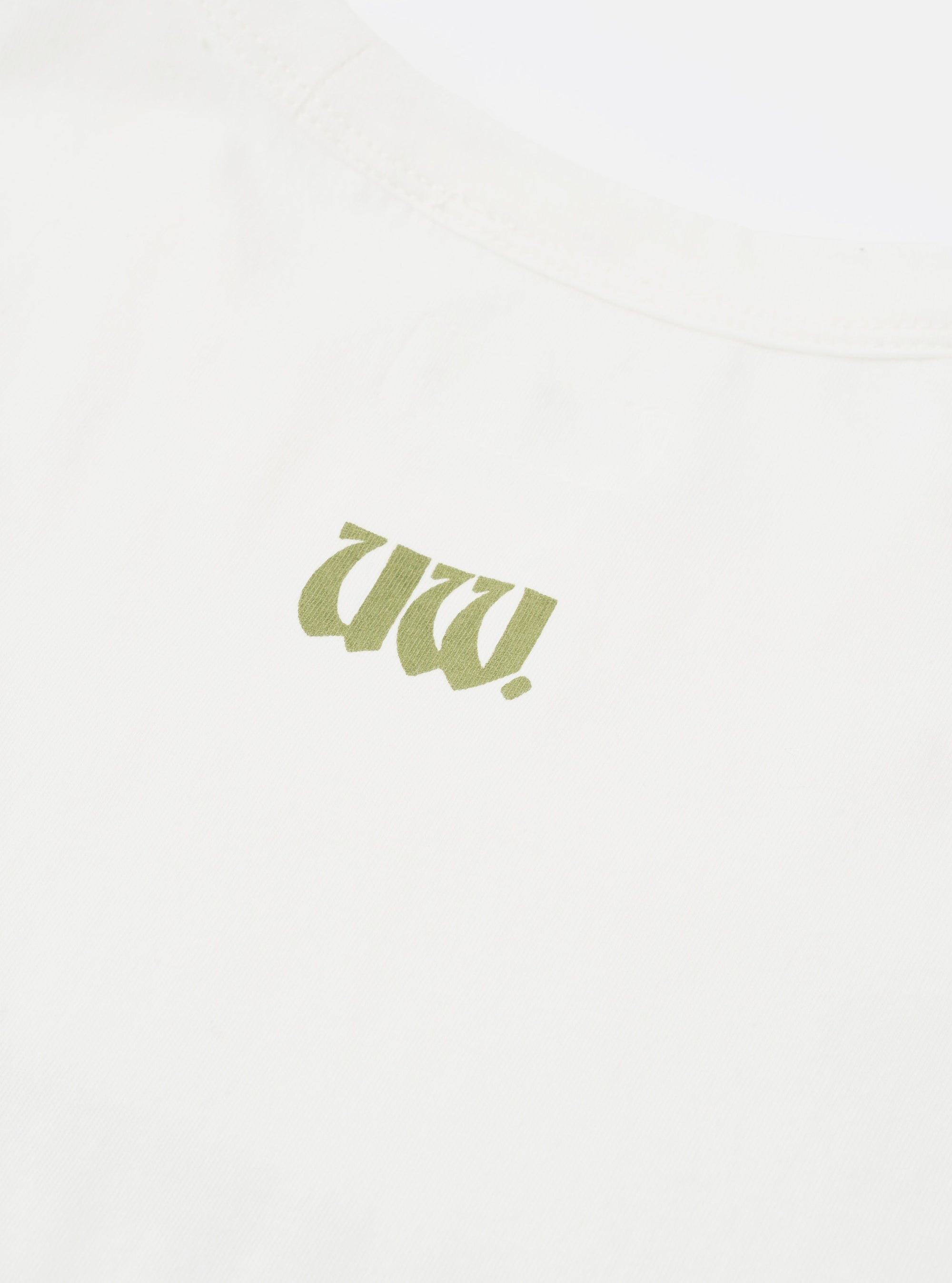 Close-up flat lay of an ecru single jersey t-shirt. A small olive green gothic-style logo print sits at centre chest. The cotton fabric texture is visible. A seam is partially visible at the top of the frame. Plain white surface background.