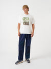 A model wearing the Universal Works Ecru Single Jersey UW Print Core Tee, shown from the front. The off-white short-sleeve cotton t-shirt features a traditional crew neckline and chest graphic print against a plain neutral backdrop.