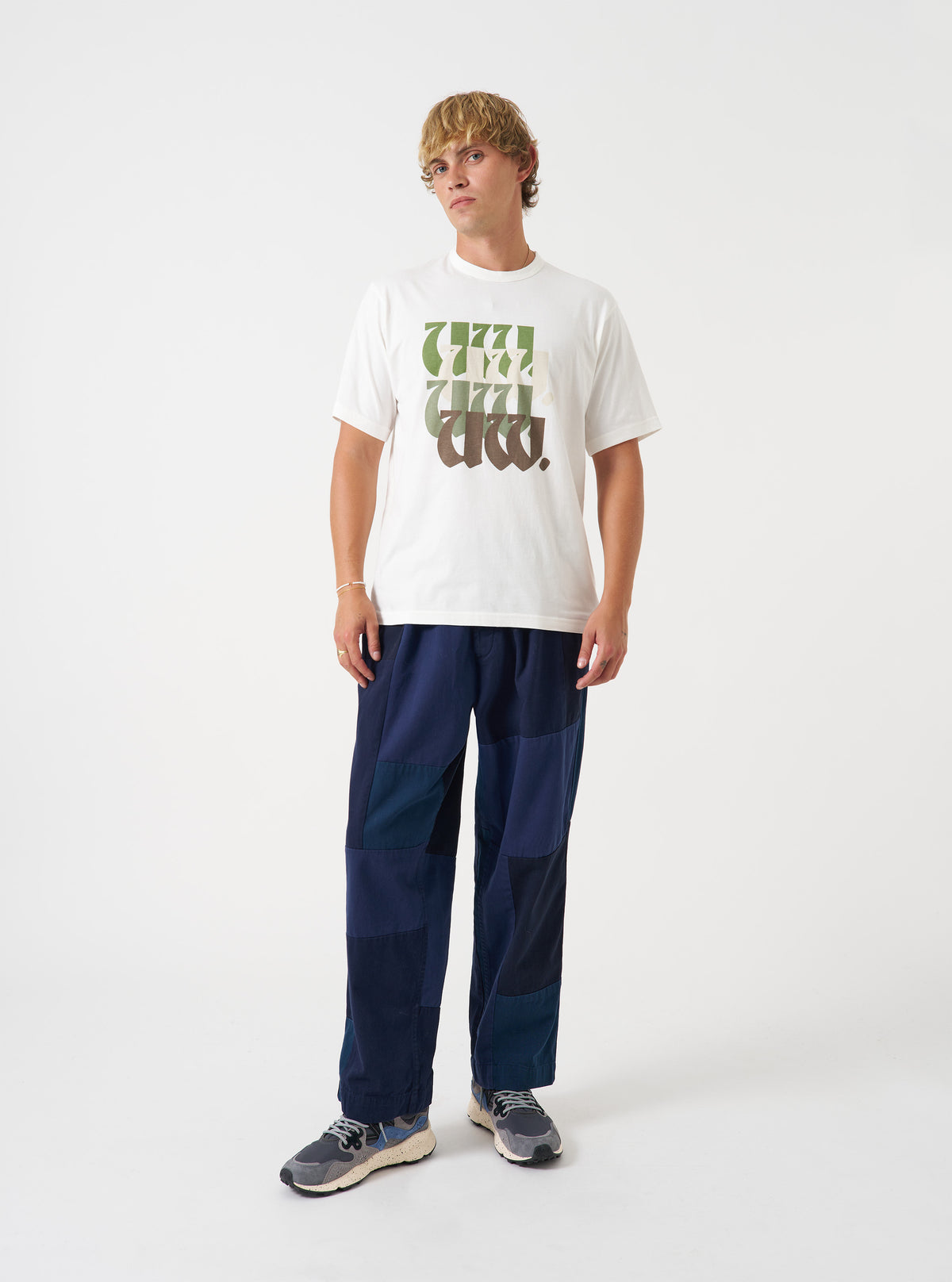 A model wearing the Universal Works Ecru Single Jersey UW Print Core Tee, shown from the front. The off-white short-sleeve cotton t-shirt features a traditional crew neckline and chest graphic print against a plain neutral backdrop.