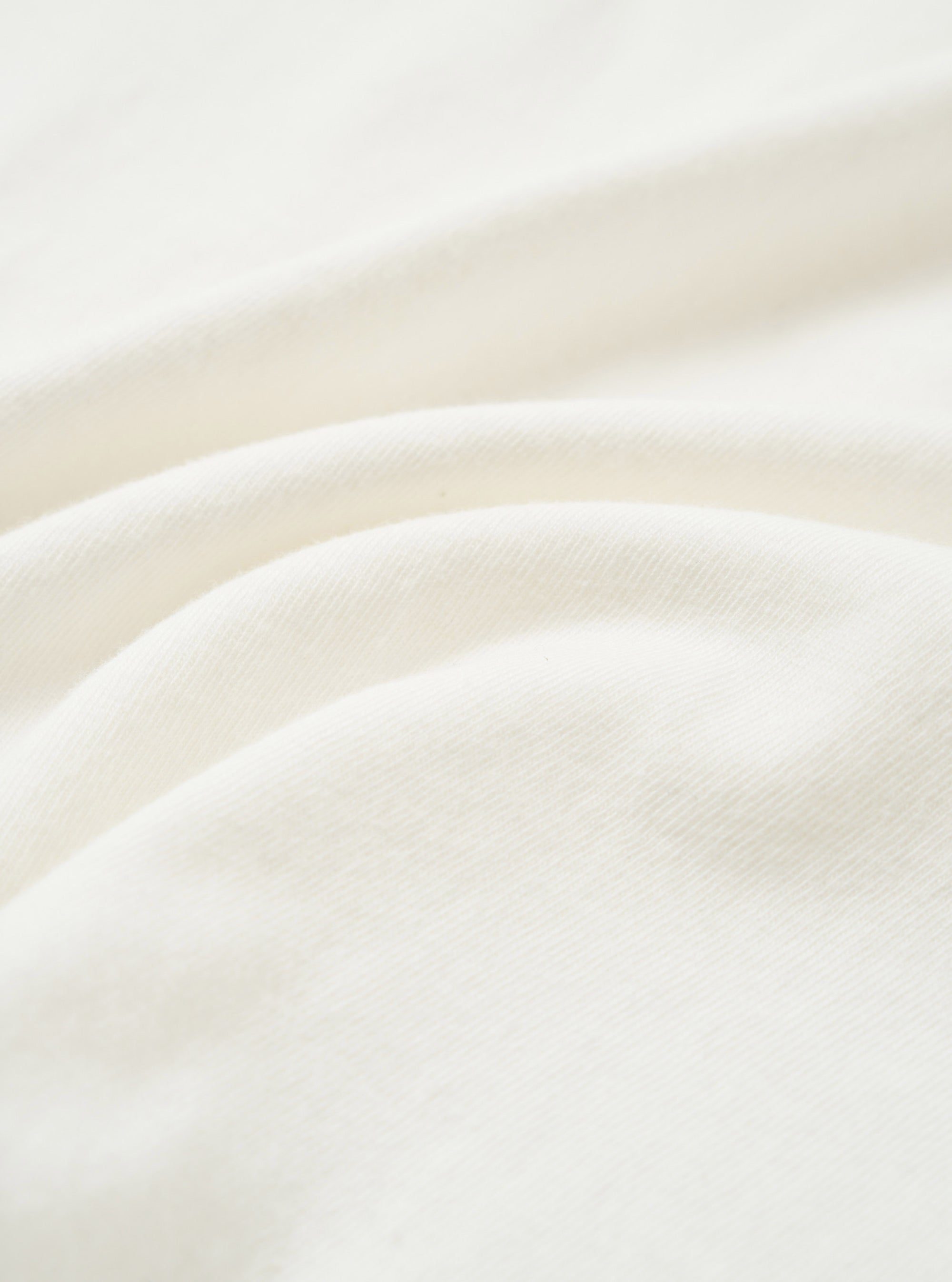 Close-up of the ecru single jersey fabric used in the Core Tee, showing a soft, fine knit texture with natural drape and gentle folds across the frame. The off-white tone is consistent throughout. No garment details or model are visible.