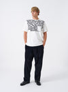 A model wearing the Universal Works Ecru Single Jersey Jazz Cool Of Jazz Core Pocket Tee, captured from the front. The light ecru short-sleeve t-shirt shows a chest pocket graphic, crew neck, and cotton finish against a plain backdrop.