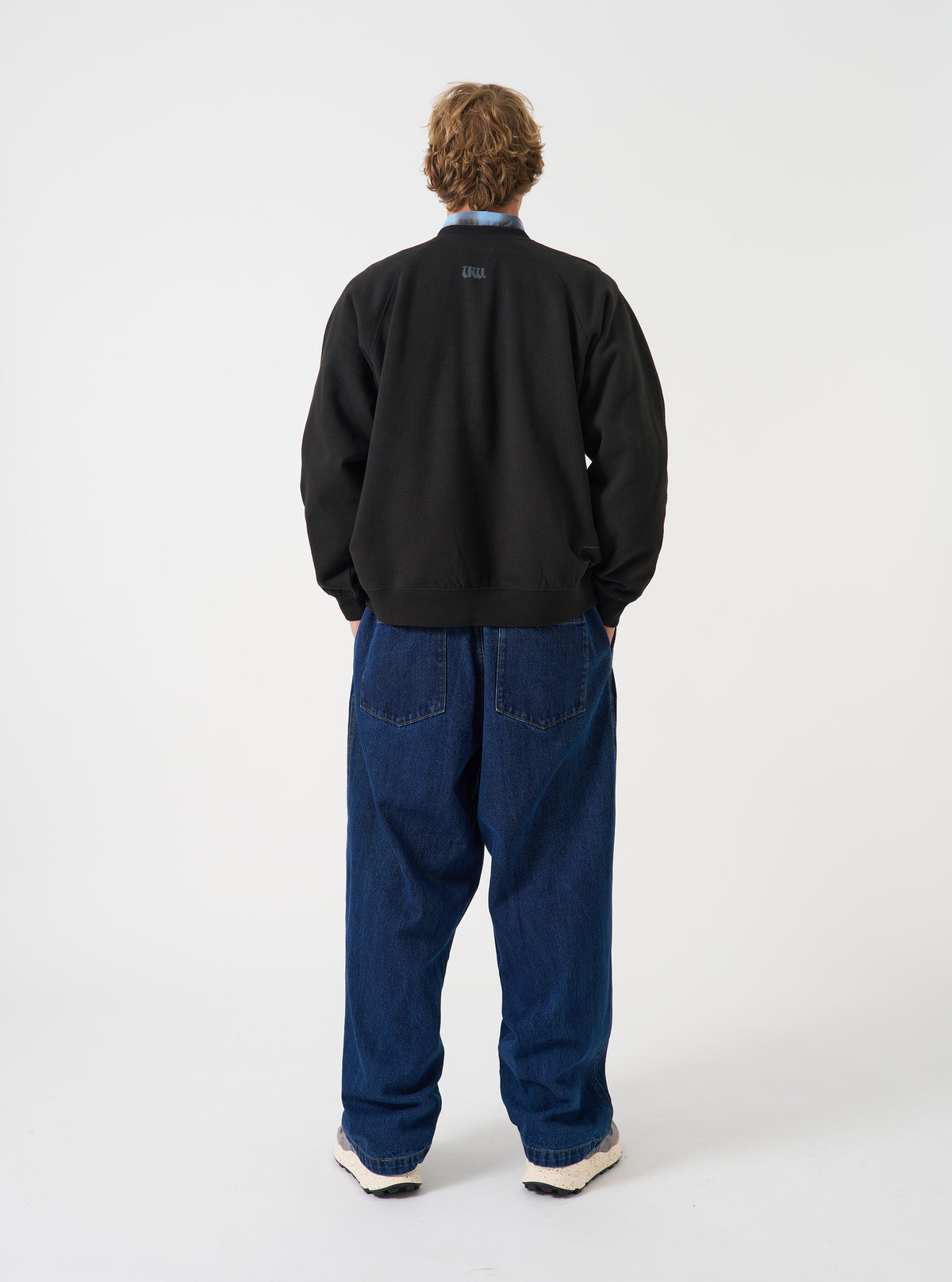 A rear view of a model wearing the Universal Works Black Brush Back Sweat UW UW Print Raglan Sweatshirt. The image highlights the raglan sleeve seams, ribbed hemline, and relaxed back fit of the black sweatshirt on a plain background.
