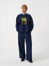 A model wearing the Universal Works Navy Brush Back Sweat UW UW Print Raglan Sweatshirt, captured from the front. The navy blue fleece-backed cotton sweatshirt displays raglan sleeves and a printed design against a plain backdrop.