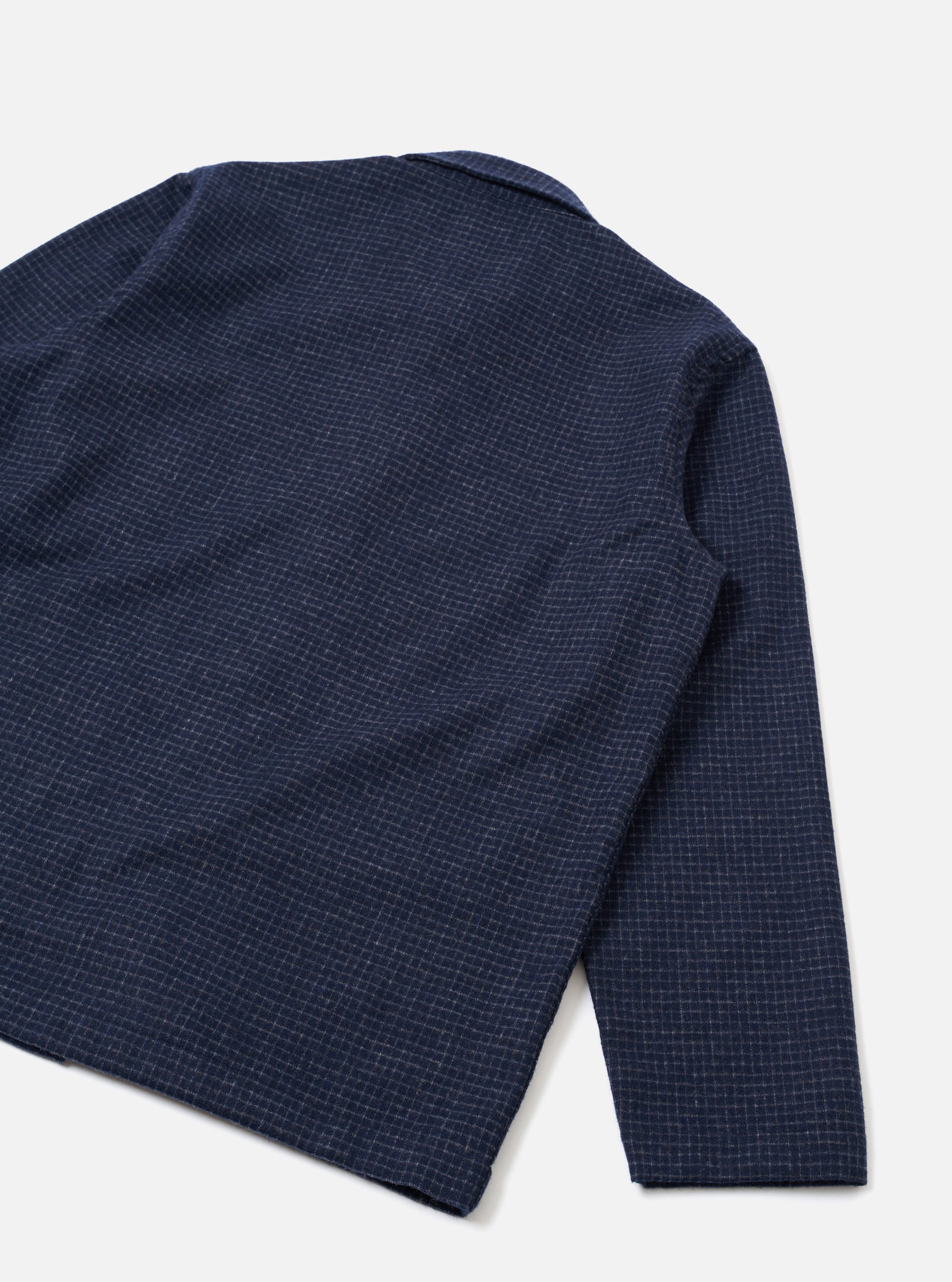 Flat lay of a navy check wool mix bakers overshirt on a light grey seamless backdrop. The shot shows the back and one sleeve at an angle. A small notched collar is visible at the top. The fabric features a fine white grid check on a navy ground with a slightly textured weave. The sleeve cuff is plain and unfinished in appearance.