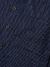 Close-up of the navy check wool mix bakers overshirt, showing three dark horn-effect four-hole buttons along the front placket and a curved patch pocket at upper right. The fabric has a fine white grid check woven into a dark navy ground with a textured, slightly nubby handle. Plain white studio backdrop.