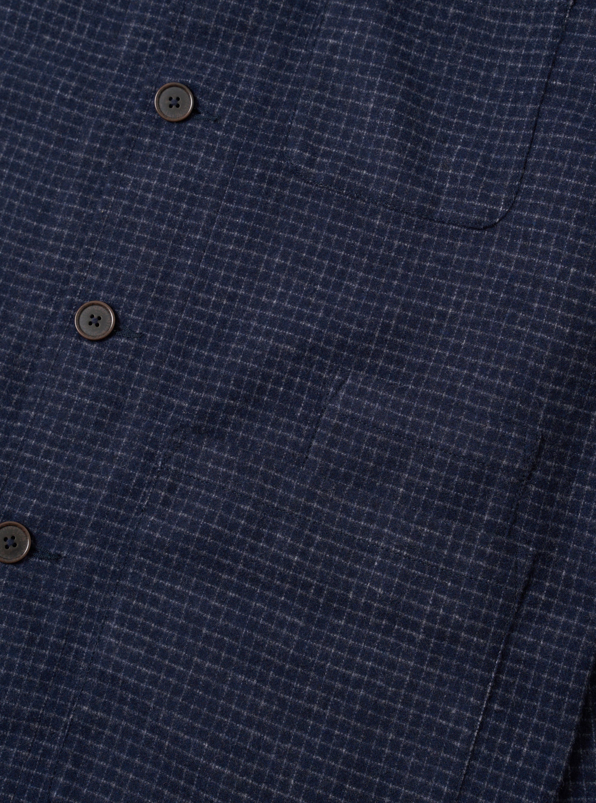 Close-up of the navy check wool mix bakers overshirt, showing three dark horn-effect four-hole buttons along the front placket and a curved patch pocket at upper right. The fabric has a fine white grid check woven into a dark navy ground with a textured, slightly nubby handle. Plain white studio backdrop.